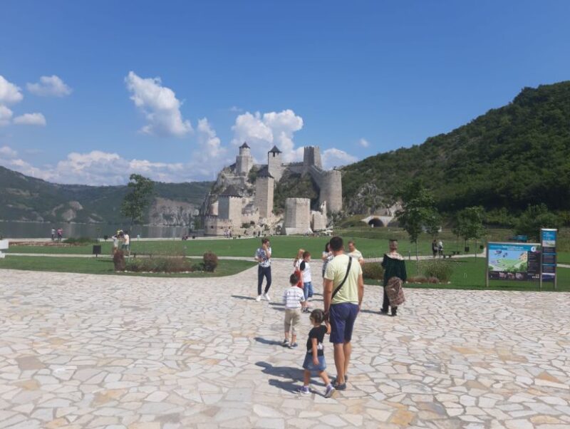 Posetioci šetaju ka Golubačkoj tvrđavi, srednjovekovnoj tvrđavi smeštenoj na obali Dunava, okruženi prelepim zelenim pejzažem i vedrim nebom.