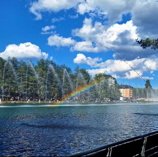 Zlatiborsko jezero sa fontanama, sunčan dan i duga iznad vode, drveće i zgrade u pozadini