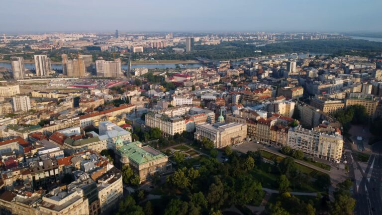 Panoramski pogled iz vazduha na centar Beograda, prikazujući istorijske zgrade, moderne građevine i ušće Save i Dunava u pozadini, pod vedrim nebom.