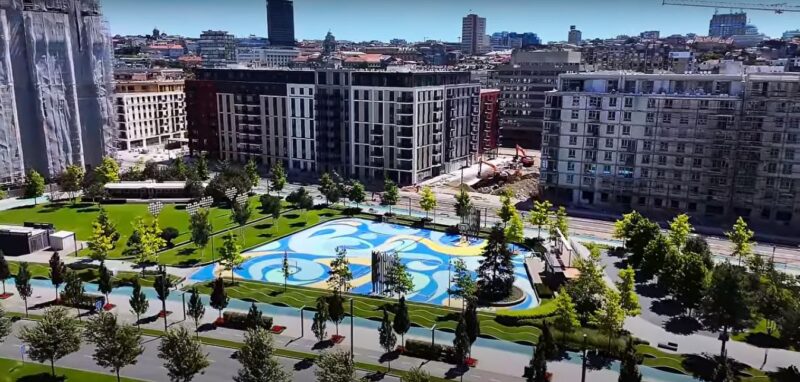 Park u Beogradu na vodi sa modernim zgradama u pozadini, zelenim površinama i umetnički dizajniranim igralištem, tokom sunčanog dana.