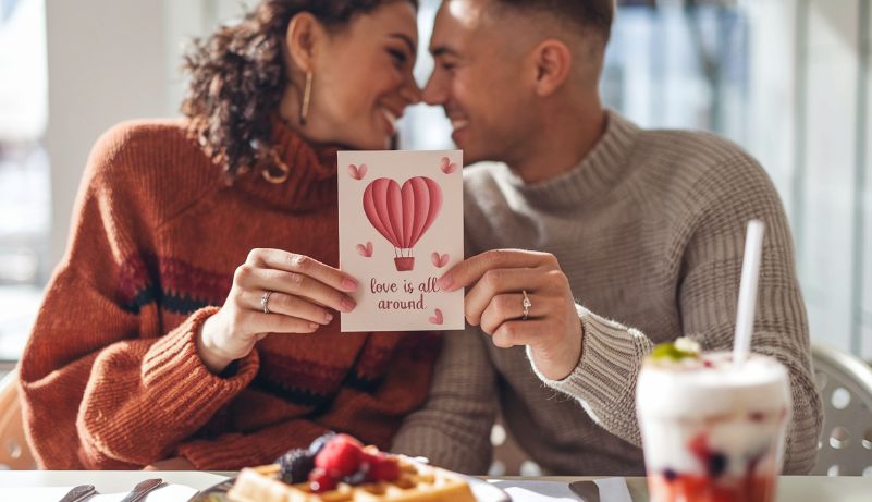 Zaljubljeni par u kafiću drži romantičnu čestitku sa natpisom „Love is all around“, dok ispred njih stoje vafli sa voćem i osvežavajući napitak.